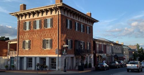 Historic brick building with blue shutters, surrounded by shops and cars under a clear sky.