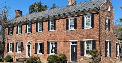 A two-story brick house with black shutters and a slate roof, surrounded by greenery under a clear blue sky.