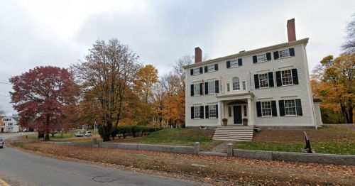 A large white historic house with black shutters, surrounded by autumn trees, along a quiet road.