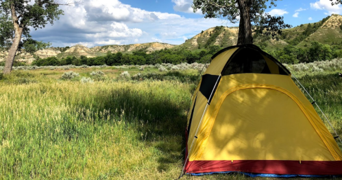 A yellow tent set up in a grassy field, surrounded by trees and hills under a partly cloudy sky.