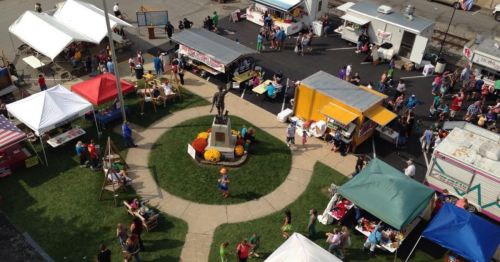 Aerial view of a bustling outdoor market with food trucks, tents, and a statue surrounded by people and colorful decorations.