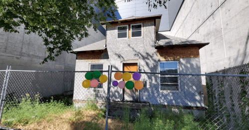 A weathered house surrounded by a chain-link fence, decorated with colorful balloons, set against a concrete backdrop.