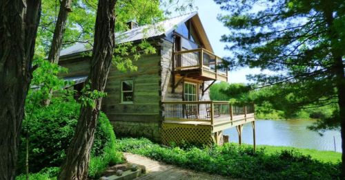 A rustic wooden cabin with a deck overlooking a serene pond, surrounded by lush greenery and trees.