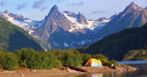 A vibrant yellow tent by a serene lake, surrounded by lush greenery and majestic snow-capped mountains in the background.