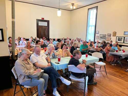 A large audience sits in a room, attentively listening to a speaker, with tables and chairs arranged for the event.