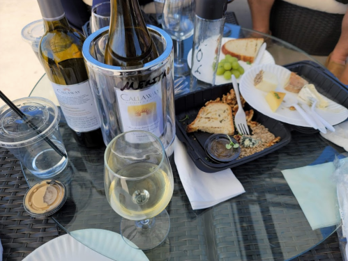 A table with wine bottles, a glass of white wine, and a platter of bread, cheese, and grapes.