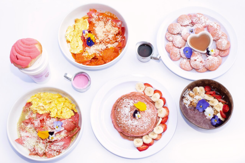 A colorful breakfast spread featuring pancakes, eggs, pastries, and a pink dessert, garnished with flowers.