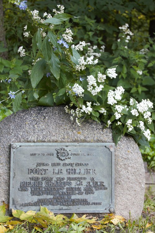 A historical plaque at Fort Le Huiller, surrounded by greenery and white flowers, commemorating its establishment.