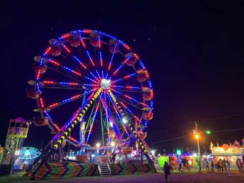 A brightly lit Ferris wheel at night, surrounded by colorful carnival attractions and people enjoying the festivities.