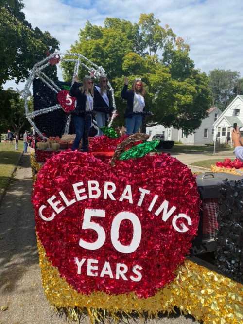 A festive parade float decorated with an oversized apple and the text "Celebrating 50 Years," featuring people waving.