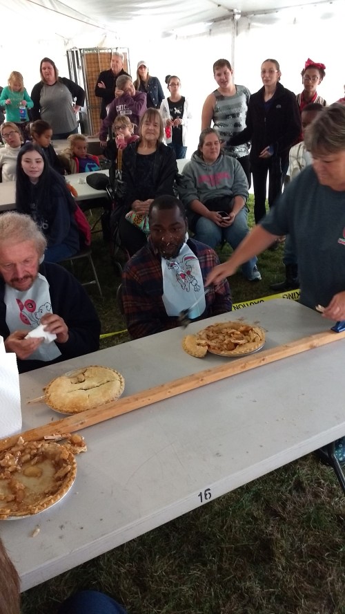 A pie-eating contest with participants at a table, surrounded by an audience in a tent. Excitement fills the air.