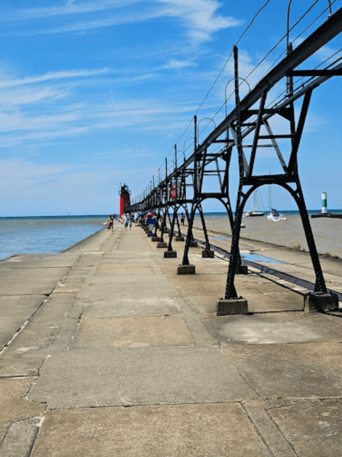 A long pier extends into the water, with a lighthouse in the distance and people walking along the path under a blue sky.