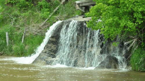 A small waterfall cascades over rocks into a brown river, surrounded by lush green foliage.
