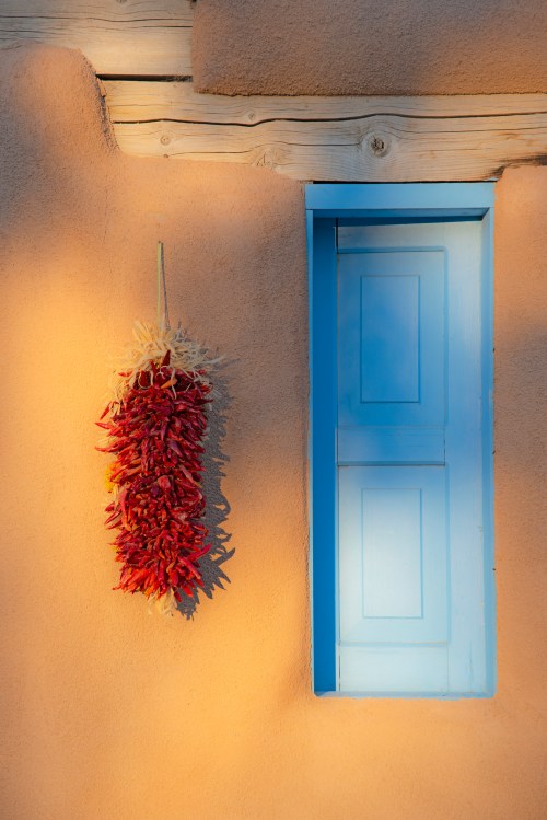 A blue window on a warm, adobe wall, with a bundle of red chilies hanging nearby.