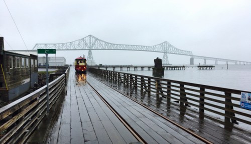 A train on wooden tracks near a foggy waterfront, with a large bridge in the background and rain on the ground.