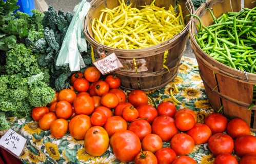 Baskets of green and yellow beans beside fresh tomatoes and kale on a floral tablecloth at a farmers market.