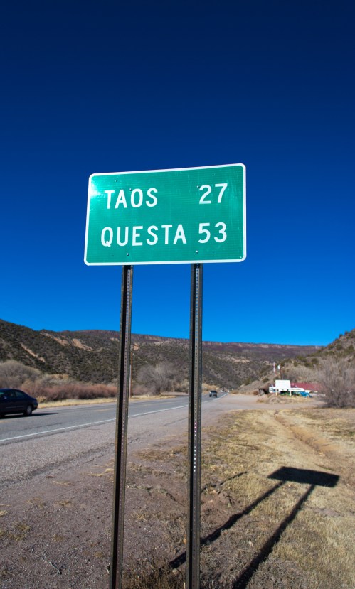 Green road sign indicating distances to Taos (27 miles) and Questa (53 miles) against a clear blue sky.