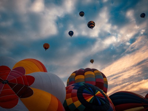 Colorful hot air balloons ascend against a dramatic sky, with clouds and vibrant patterns creating a festive atmosphere.
