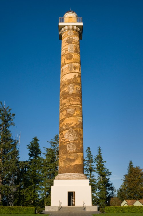 A tall, decorated lighthouse tower with intricate carvings, surrounded by trees and a clear blue sky.