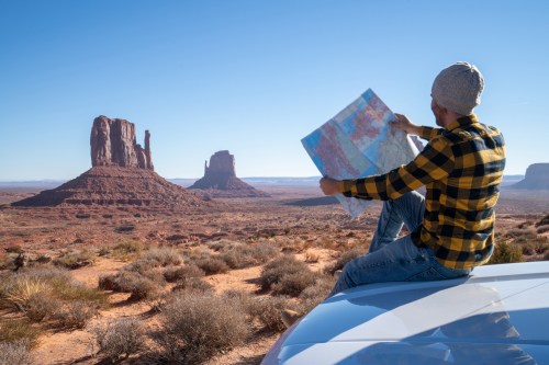 A person in a beanie and flannel shirt sits on a car, studying a map with Monument Valley in the background.