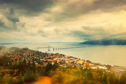 A scenic view of a coastal town with a bridge over water, surrounded by trees and cloudy skies.