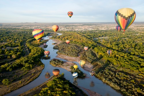 Aerial view of colorful hot air balloons over a winding river surrounded by lush greenery and a distant cityscape.