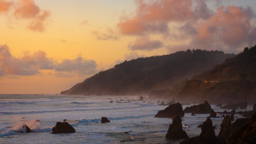 Sunset over a rugged coastline with waves crashing against rocky formations and mist rising from the shore.