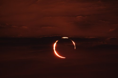 A dramatic sunset with a crescent shape of sunlight peeking through dark clouds, creating a striking silhouette.