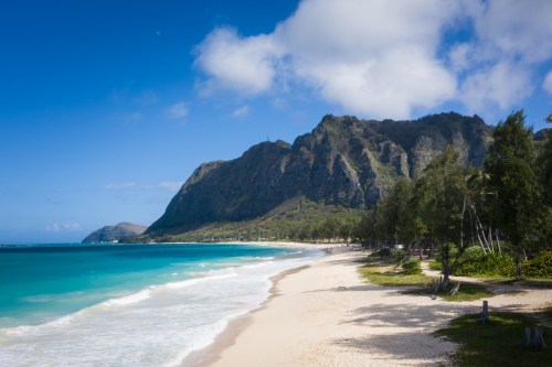 A serene beach with turquoise waters, soft sand, and lush mountains under a blue sky with scattered clouds.