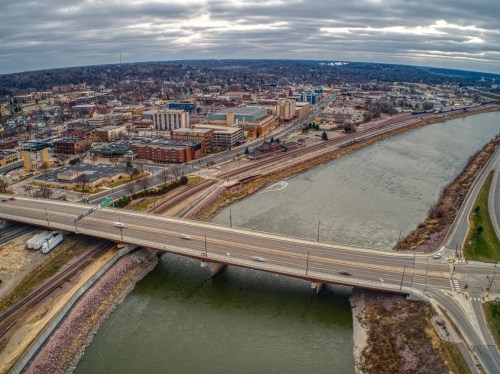 Aerial view of a city with a river, bridge, and roads, under a cloudy sky. Urban landscape with buildings in the background.