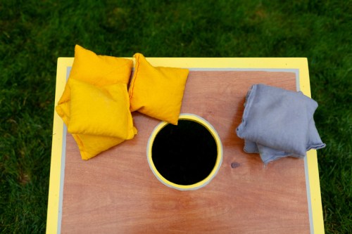 A wooden game board with a hole in the center, featuring yellow and gray bean bags stacked beside it on green grass.