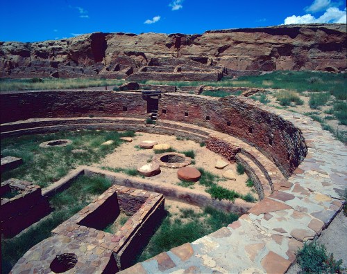 Ancient circular ruins surrounded by grass and rocky cliffs under a bright blue sky.