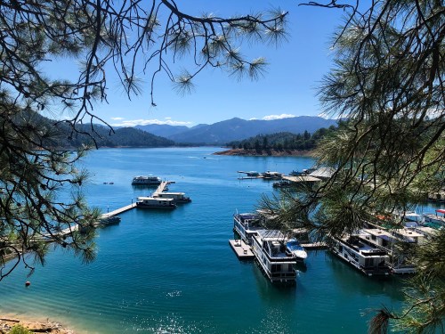A serene lake scene with boats docked, surrounded by mountains and trees under a clear blue sky.