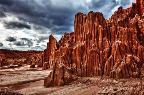 Dramatic rock formations rise against a cloudy sky, showcasing rich red and orange hues in a rugged landscape.