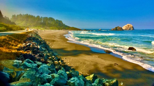 A serene beach scene with gentle waves, rocky shore, and lush trees in the background under a clear blue sky.