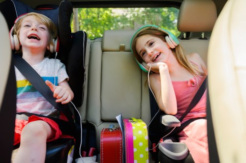 Two children in car seats, wearing headphones, smiling and enjoying music during a car ride.