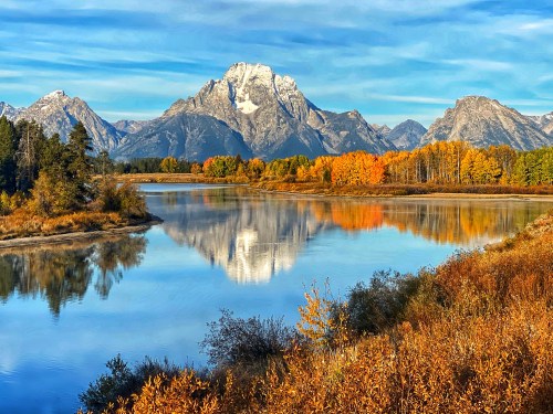 Scenic view of mountains reflecting in a calm river, surrounded by autumn foliage and a clear blue sky.