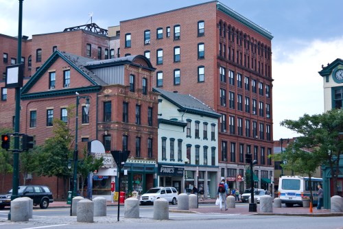 Historic buildings line a city street, featuring a mix of architectural styles and a cloudy sky overhead.