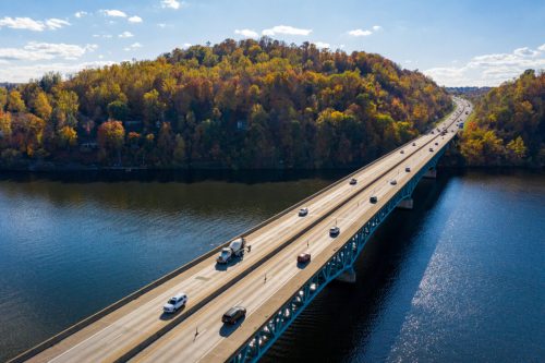 Aerial view of a highway bridge over a river, surrounded by autumn-colored trees and clear blue skies.