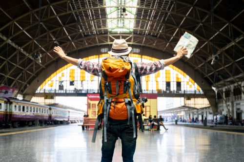 A traveler with a backpack stands in a train station, arms outstretched, holding a map, ready for adventure.