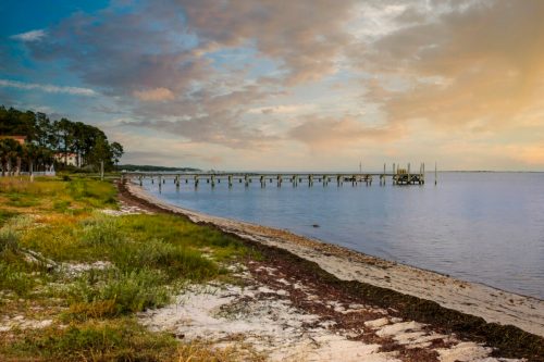 A serene waterfront scene with a wooden pier extending into calm waters, surrounded by lush greenery and a cloudy sky.
