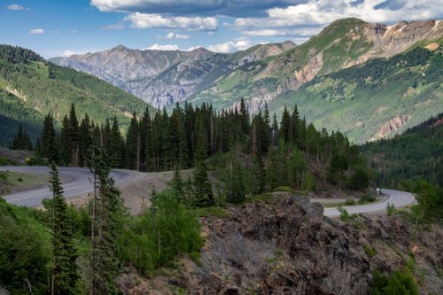 A winding road through lush green forests, with majestic mountains in the background under a partly cloudy sky.