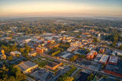 Aerial view of a small town at sunrise, showcasing buildings, streets, and greenery under a misty sky.