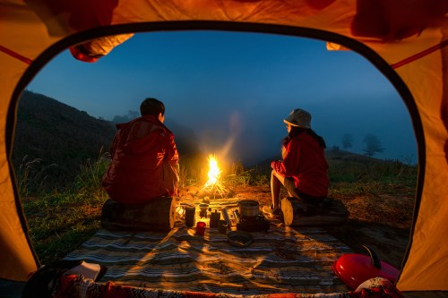 Two people sit by a campfire inside a tent, surrounded by nature at dusk, enjoying a cozy outdoor experience.
