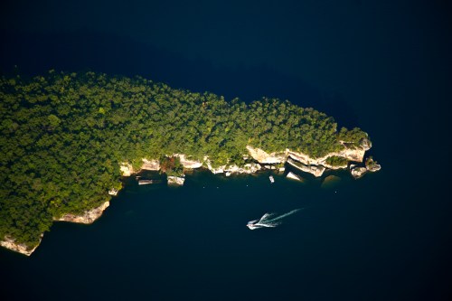 Aerial view of a lush green island with rocky cliffs, surrounded by dark blue water and a small boat nearby.