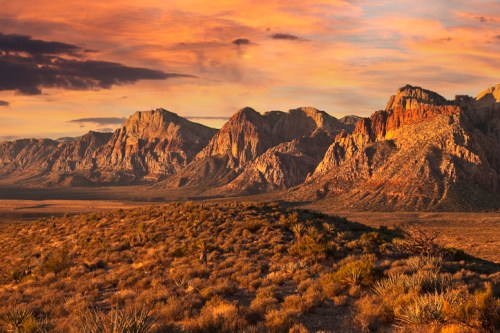 Sunset over rugged mountains with vibrant orange and purple skies, highlighting the desert landscape below.