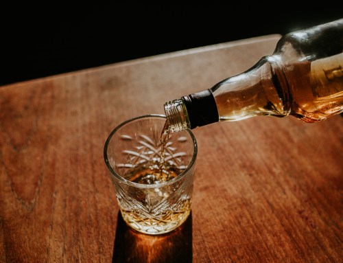 A bottle pours whiskey into a crystal glass on a wooden surface, illuminated by soft sunlight.
