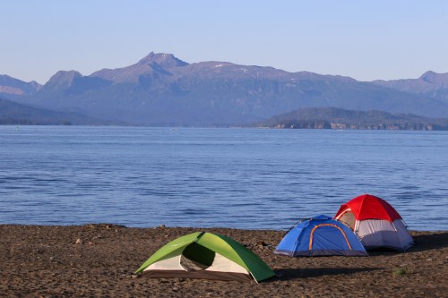 Three colorful tents on a sandy beach by a calm lake, with mountains in the background under a clear blue sky.