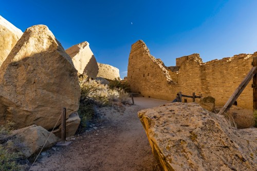 Ancient stone ruins under a clear blue sky, with rocky formations and a path leading through the site.