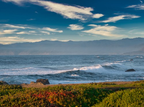 A serene coastal scene with waves crashing on the shore, framed by green vegetation and distant mountains under a blue sky.